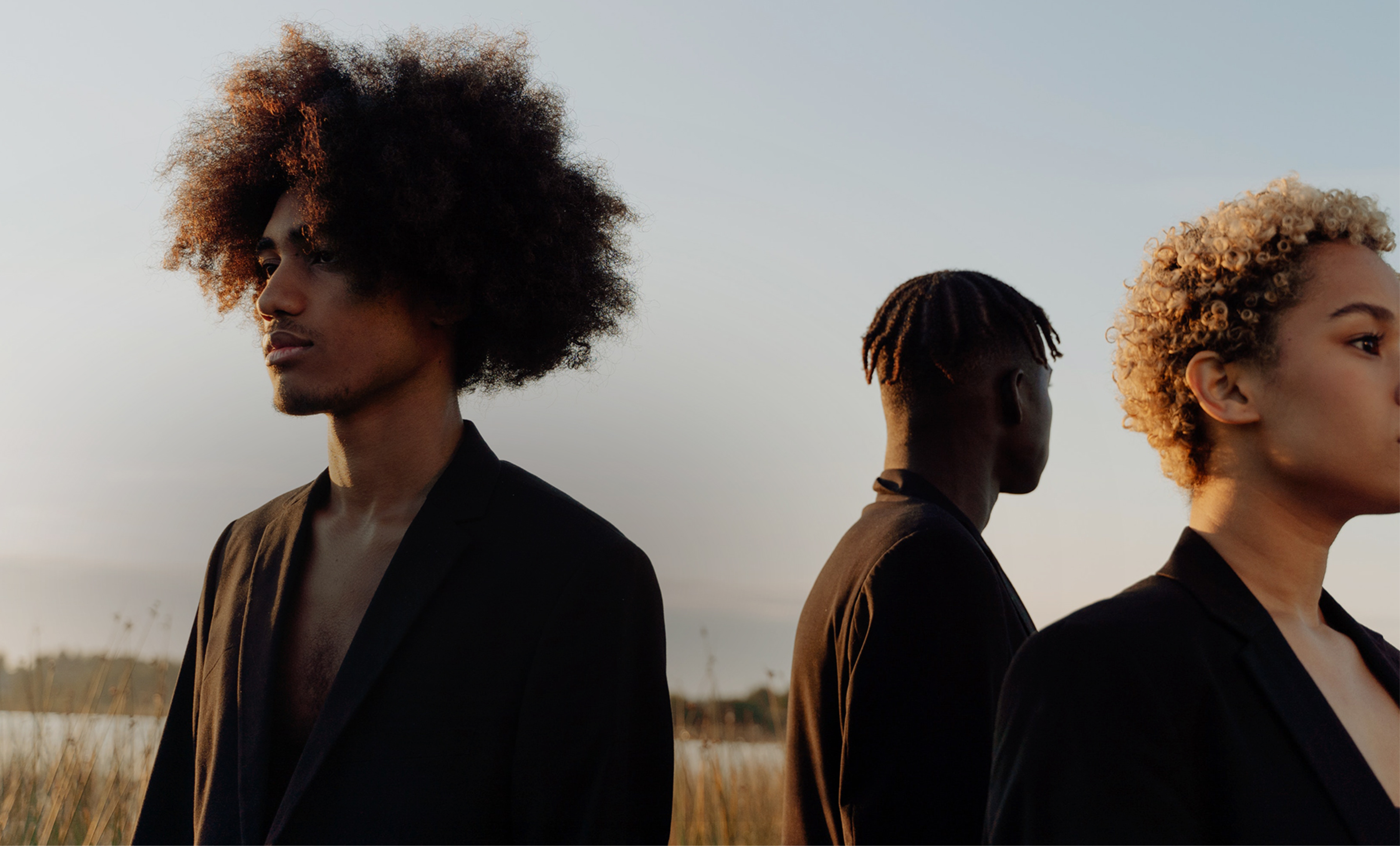 Three people with diverse hairstyles wearing dark clothing against a sunset background